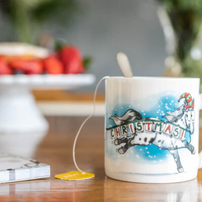 Image of Emily Cole's Happy Christmas Fine Bone China Mug, showcasing a spotted pony and a 'Happy Christmas' banner against a snowy background